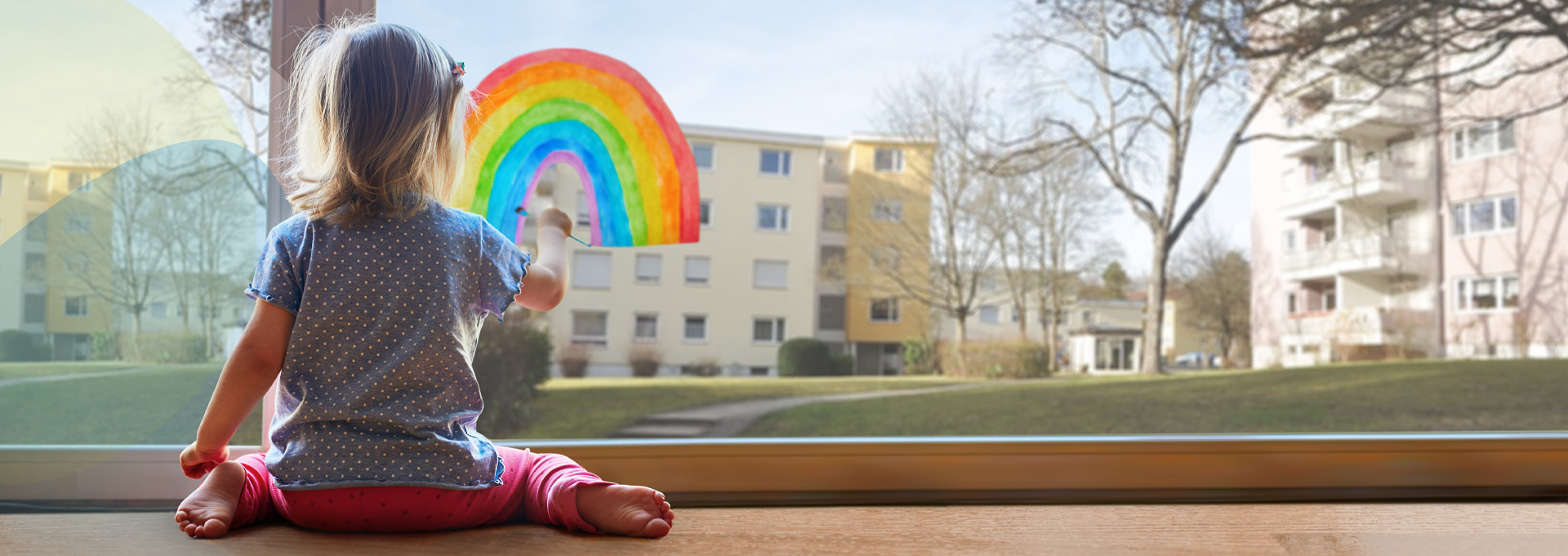 Kleines Kind auf warmem Fußboden durch Fernwärme in Böblingen. Malt einen Regenbogen an ein Terassenfenster. Hinter ihr bunte SWBB Sphären.