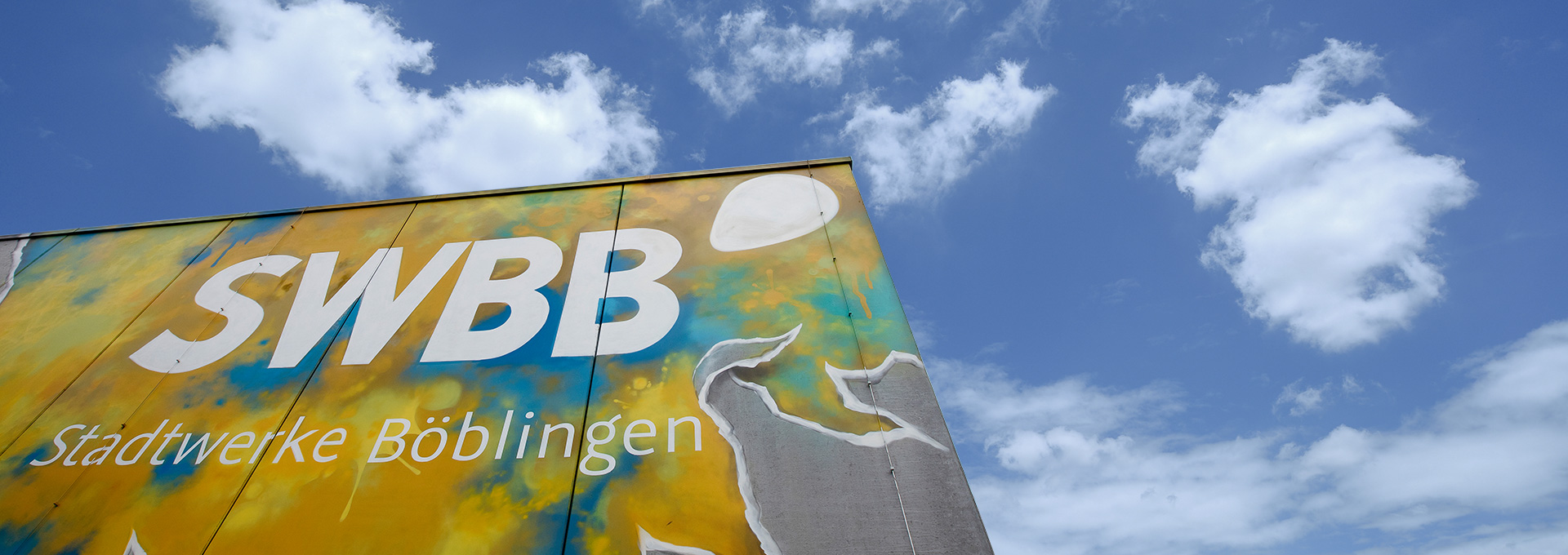 BHKW der Stadtwerke Böblingen an der Ernst-Reuter-Straße, Bild zeigt einen Ausschnitt eines Graffittis mit dem SWBB Logo und der Beschriftung Stadtwerke Böblingen auf gelb-blauem Hintergrund. Blauer Himmel mit Wolken im Hintergrund.