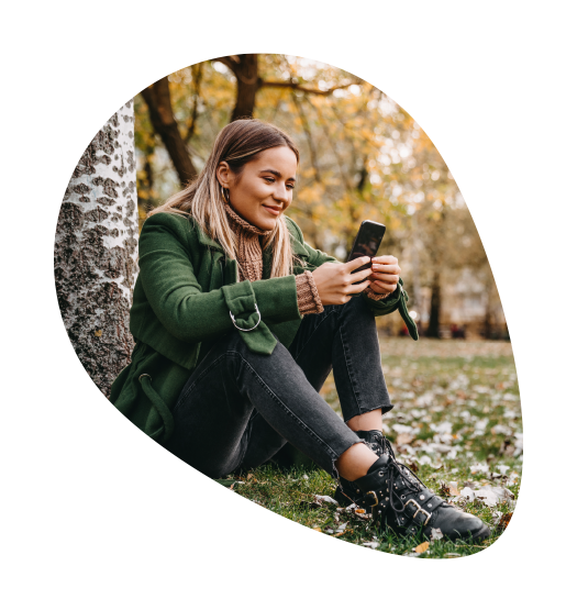 Lächelnde Frau mit grünem Mantel sitzt an einen Baum gelehnt auf einer herbstlichen Grünfläche und schaut in ihr Handy.