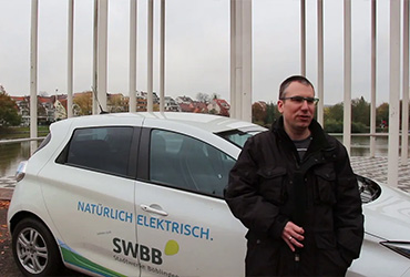 Carsten Scharfenberg gibt Interview zur E-Mobilität im Alltag. Der Mann mit Brille hält ein Mikrofon und steht vor einem E-Auto mit der Beschriftung "Natürlich elektrisch - SWBB"