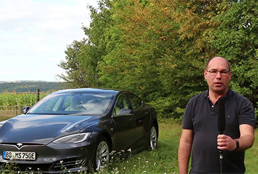 Michael Bär gibt Interview zur Reichweite und Lademöglichkeiten mit Elektroautos: Mann mit Brille und Mikrofon steht auf einer Wiese, neben ihm steht ein schwarzes E-Auto der Marke Tesla.