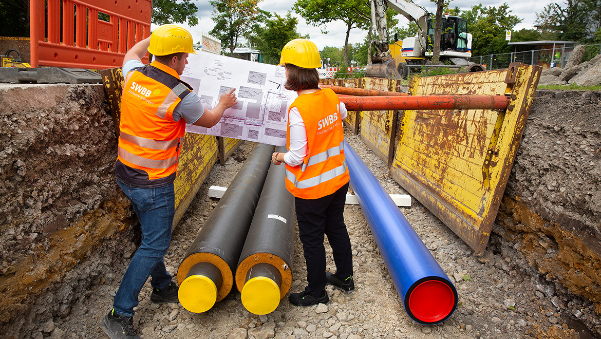 Baustelle der SWBB zur Verlegung der Fernwärmerohre: Zwei von der Kamera weggedrehte Personen in Warnwesten, die in einer Baugrube mit Rohren stehen und einen Plan betrachten.