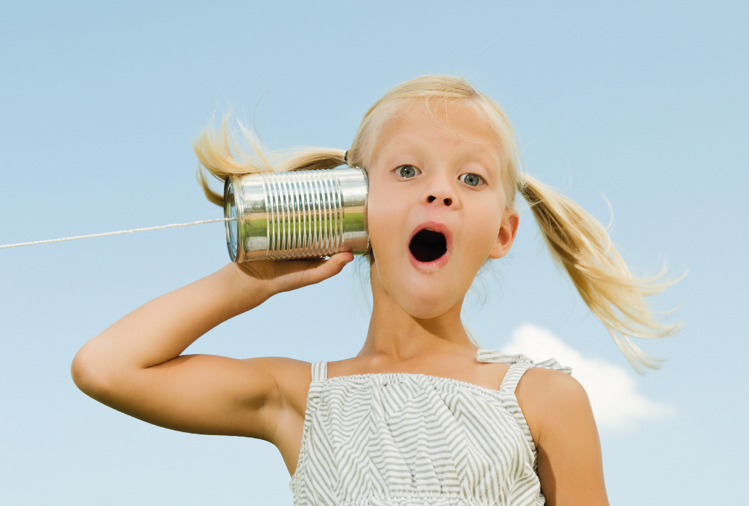 Blondes Mädchen hält sich spielerisch ein Dosentelefon an das Ohr. Im Hintergrund der blauer Himmel.